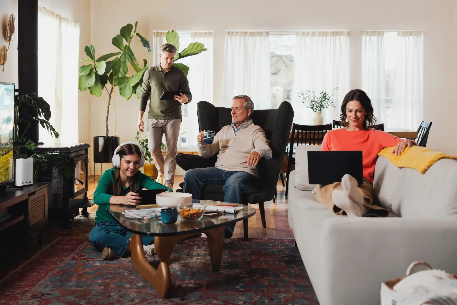 A multi-generational family uses various electronic devices including a laptop, tablets, and headphones while relaxing together in a brightly lit, modern living room.