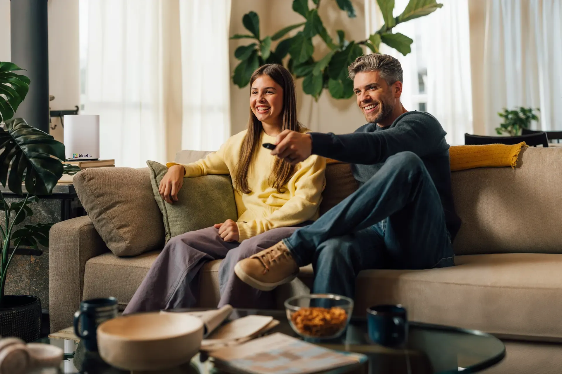A father and daughter sitting on a living room sofa, smiling and using a remote to stream content on a television.