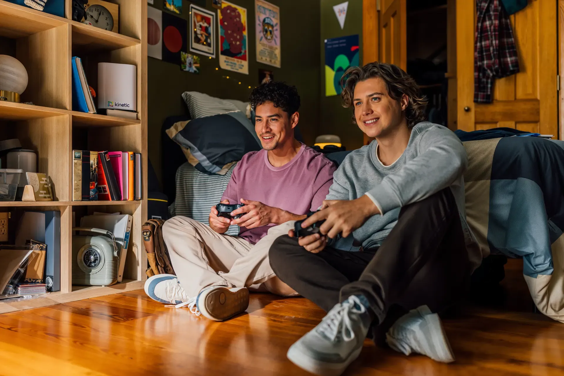 Two young men sitting on a wooden floor in a bedroom, smiling while holding controllers and playing video games.