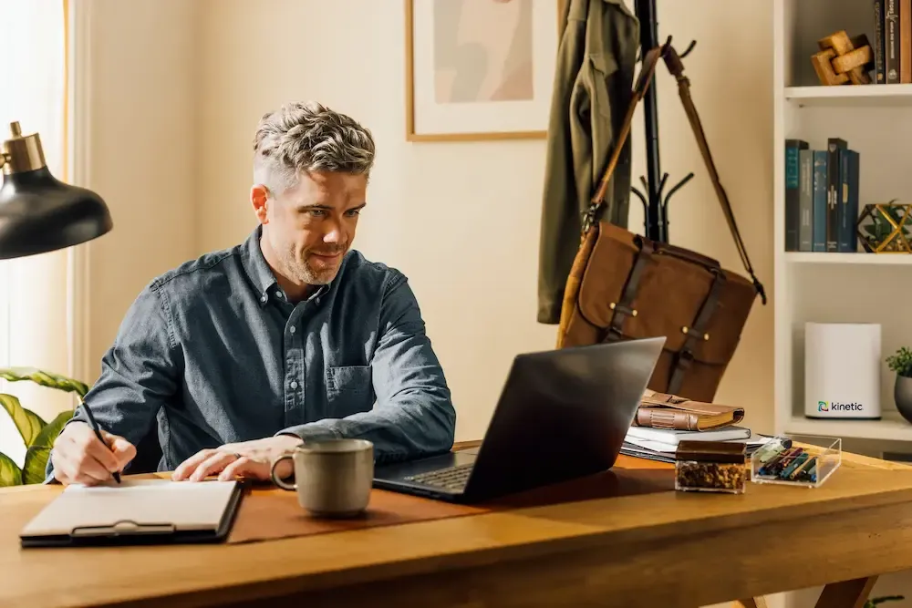 A man writes notes at a wooden desk while working on a laptop near a Kinetic Wi-Fi router