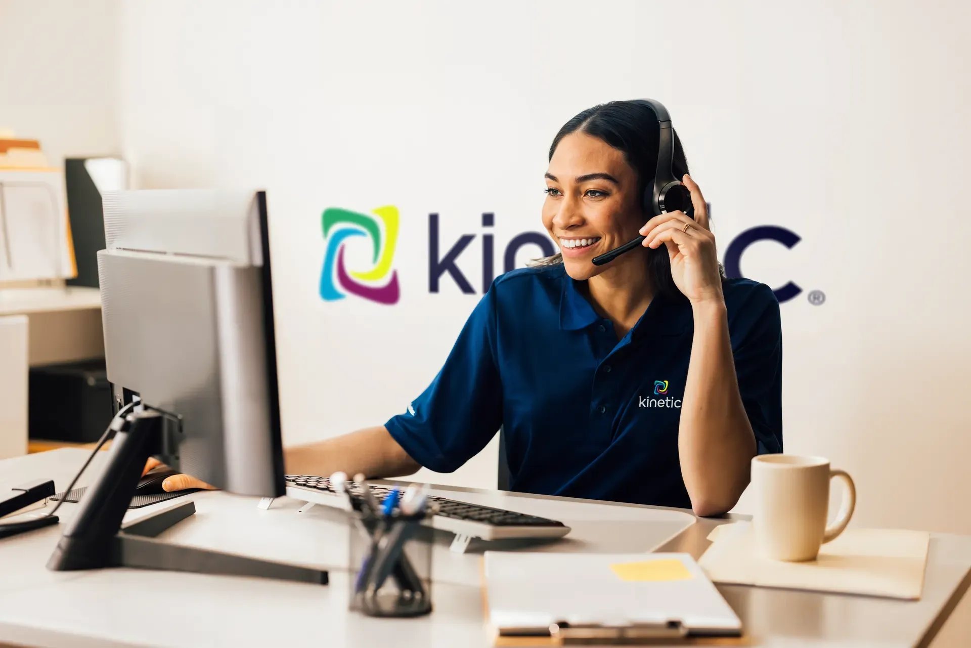 A smiling female customer service representative wearing a headset and a Kinetic branded polo shirt works at a desk with a computer.