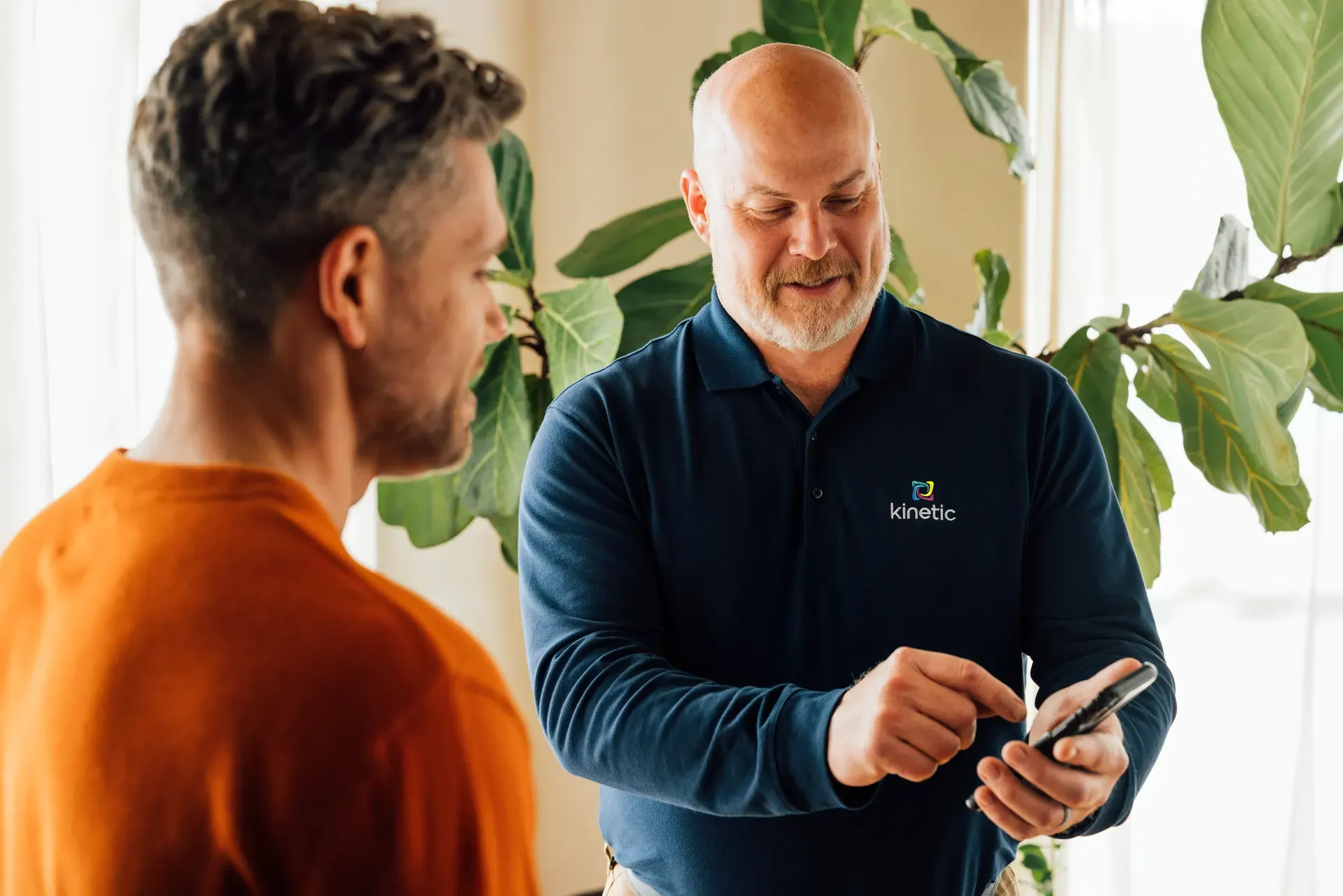 A Kinetic-branded employee shows a customer something on a smartphone beside a leafy plant.