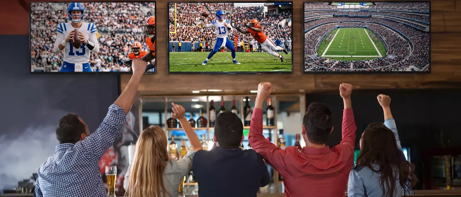A group of patrons sitting at a bar cheer with their arms raised as three large screens display live football game coverage.