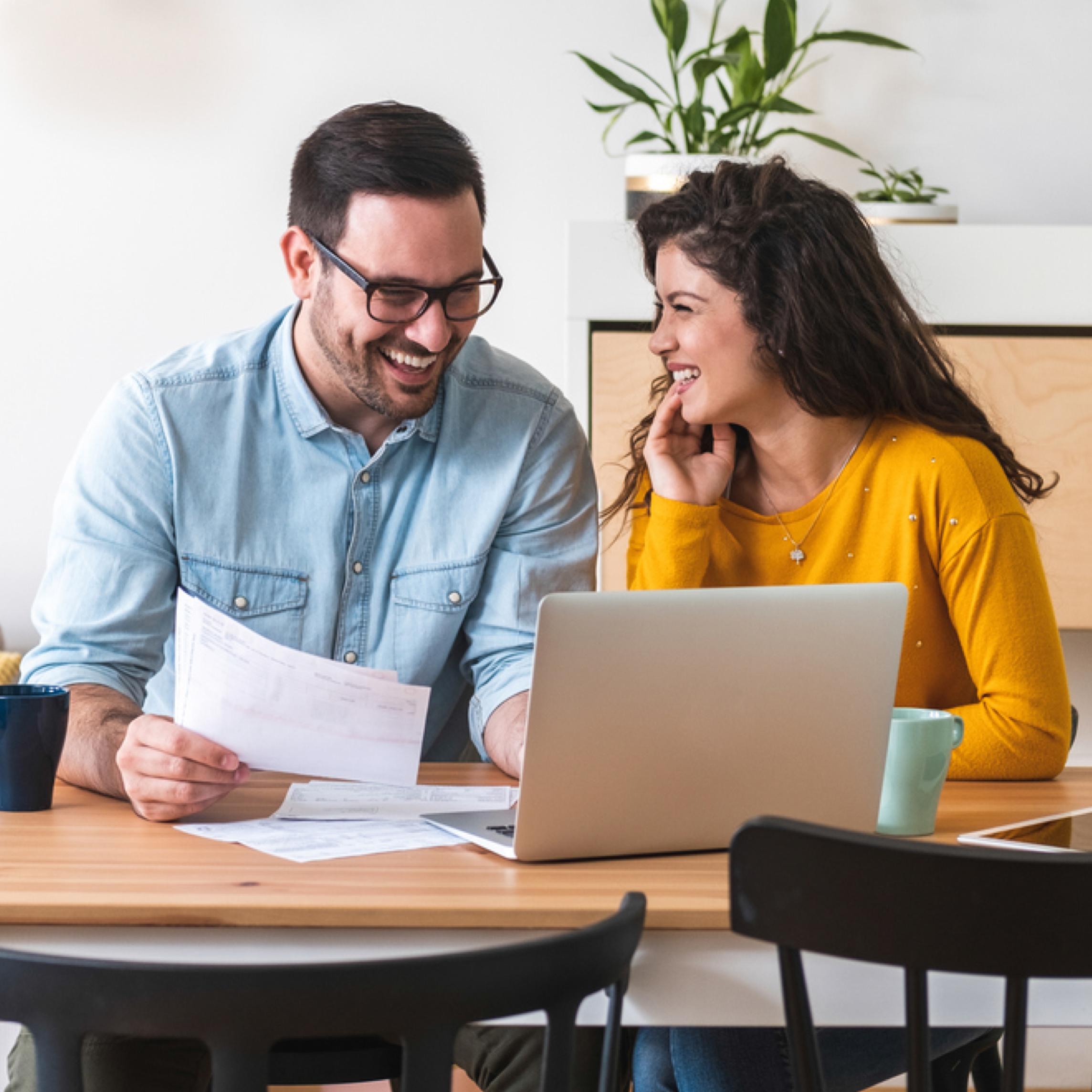 Couple sits at table and laughs together as they look at laptop and hold papers.