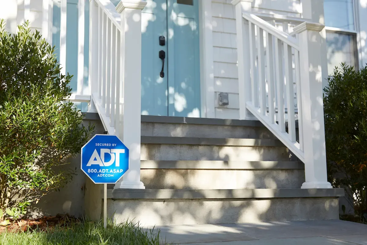 A blue ADT security yard sign stands in a garden bed next to the front steps of a white house with a blue door.