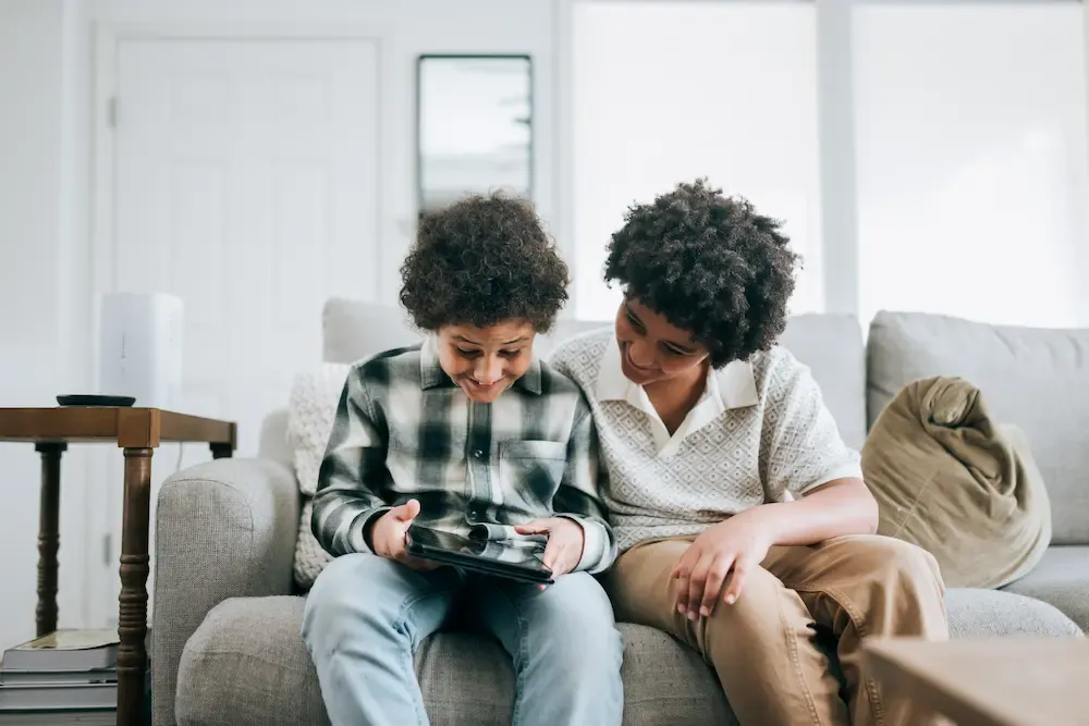 Two smiling children sit on a couch sharing a tablet in a bright living room.