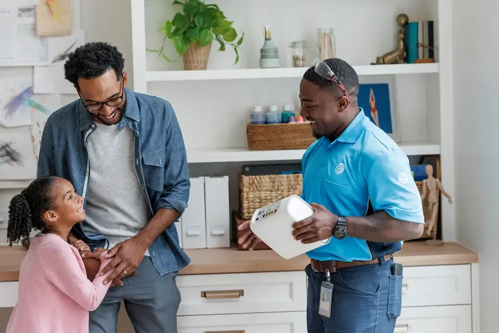 A smiling technician in a blue uniform shows an internet gateway to a man and a young girl in their home.