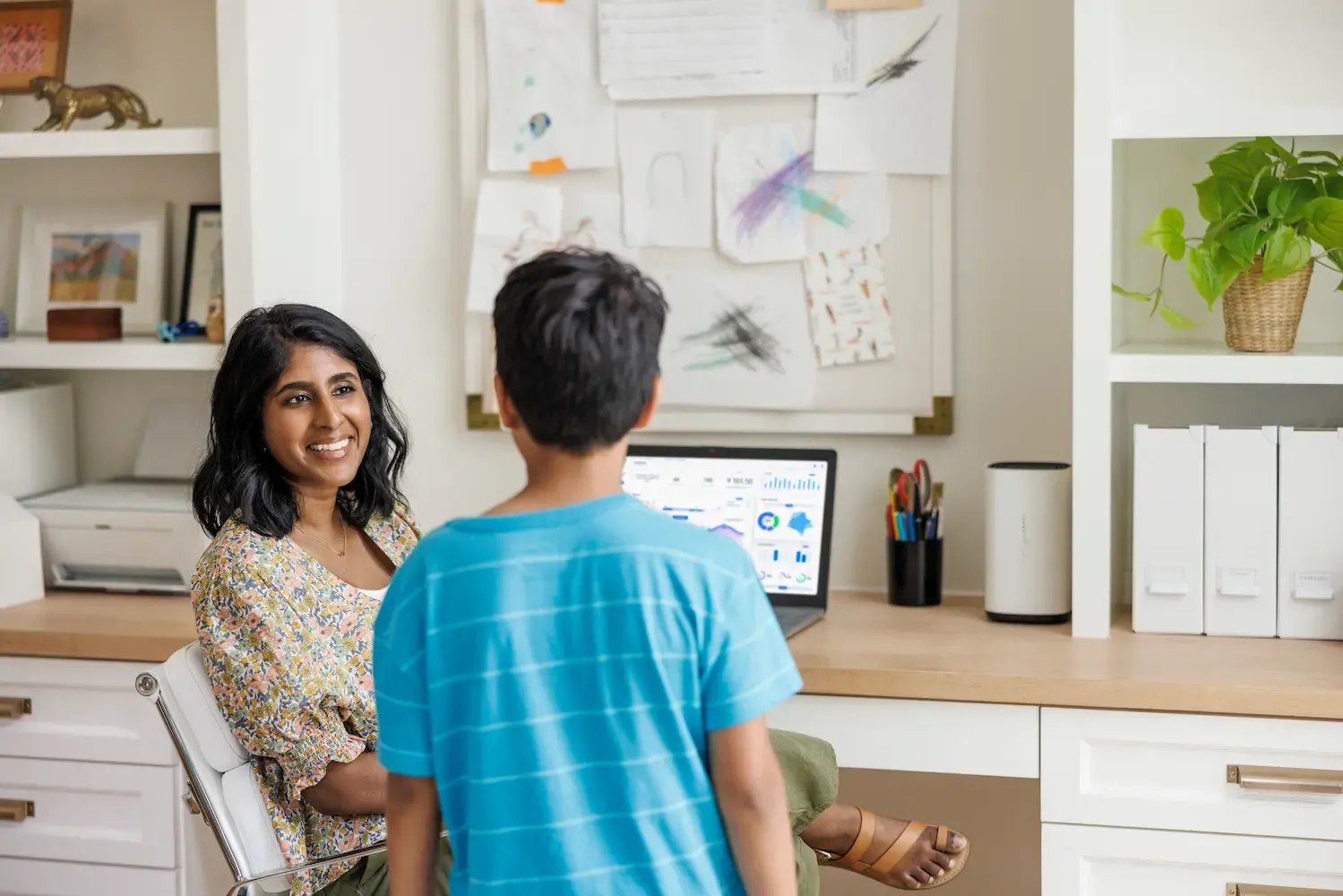A smiling woman sits at a desk in a home office and looks at a young boy standing in front of her.