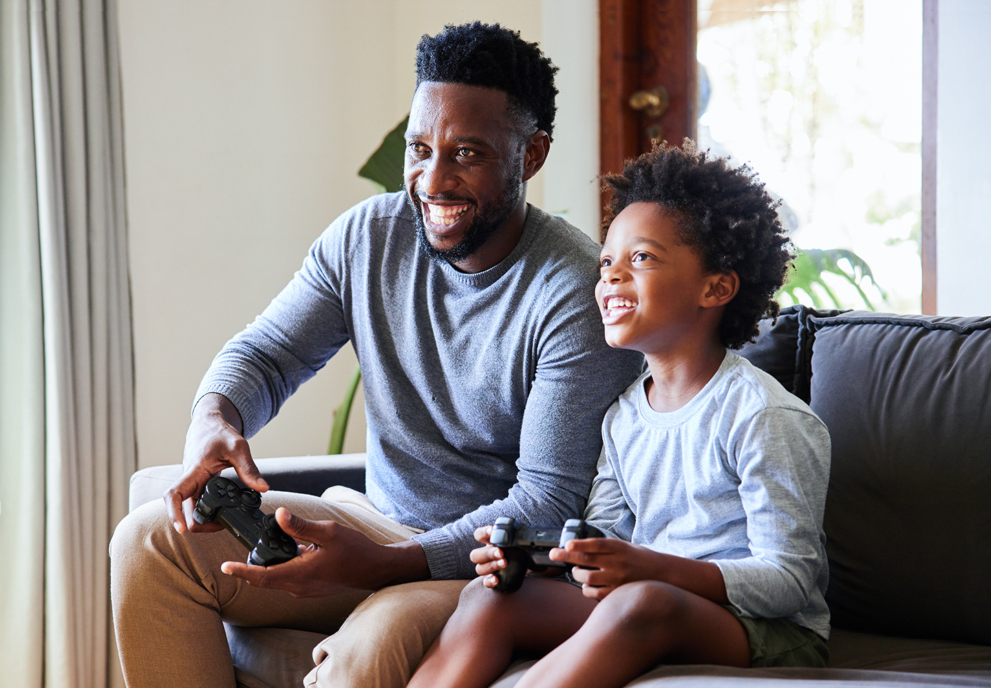 Little boy and his dad laughing and playing video games while sitting on a sofa in their living room at home