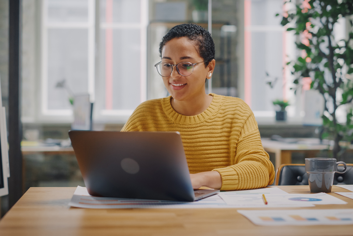Chica con gafas y suéter amarillo, buscando en internet en su computadora portátil.