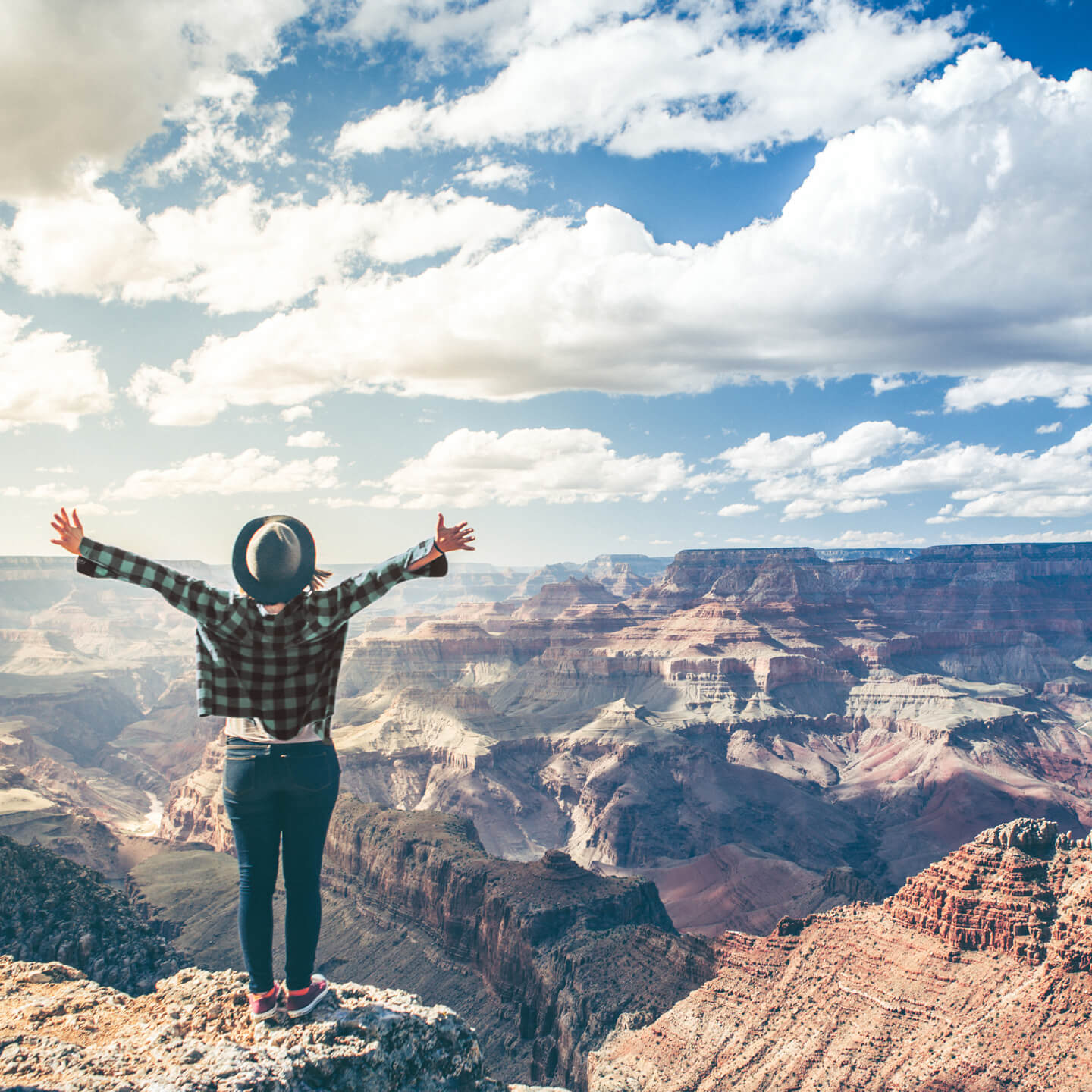 Young girl in green flannel shirt and felt hat raising arms to the sky and looking out on the Arizona side of the Grand Canyon.