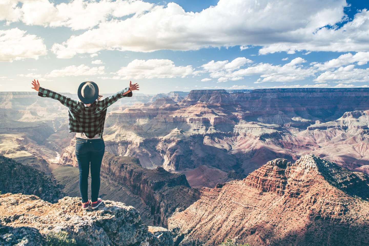 Young girl in green flannel shirt and felt hat raising arms to the sky and looking out on the Arizona side of the Grand Canyon.