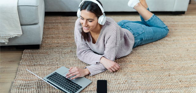 Teen girl wearing headphones laying on floor with laptop and cell phone