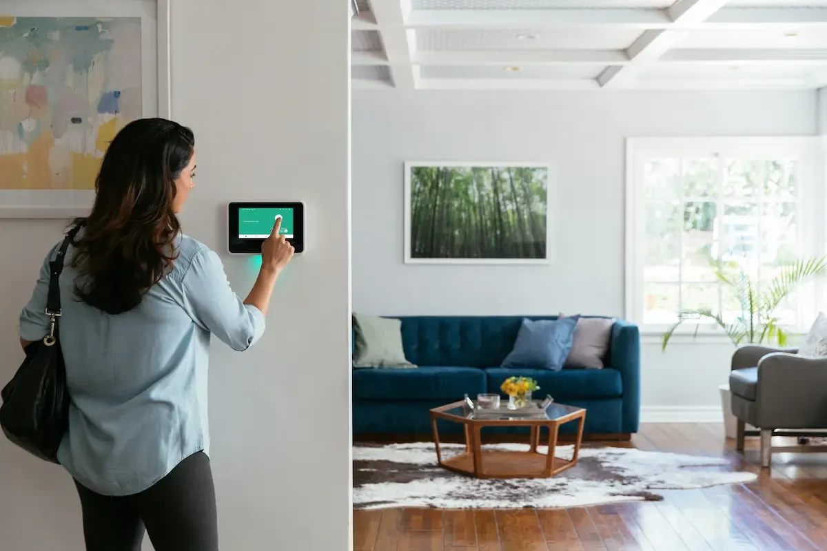 A woman with a shoulder bag interacts with a wall-mounted Vivint Smart Hub control panel in a modern living room.