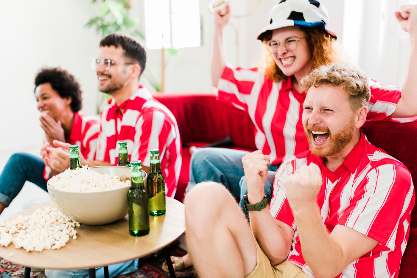 Group of four friends, watching a professional soccer match, all are wearing matching red and white horizontal stripe jerseys, and eating popcorn and drinking beers