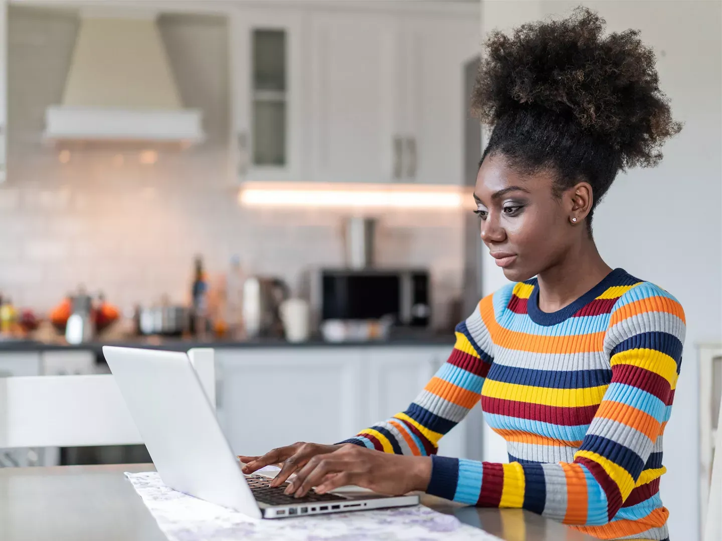 A focused woman wearing a colorful striped sweater sits at a kitchen table while typing on her laptop.