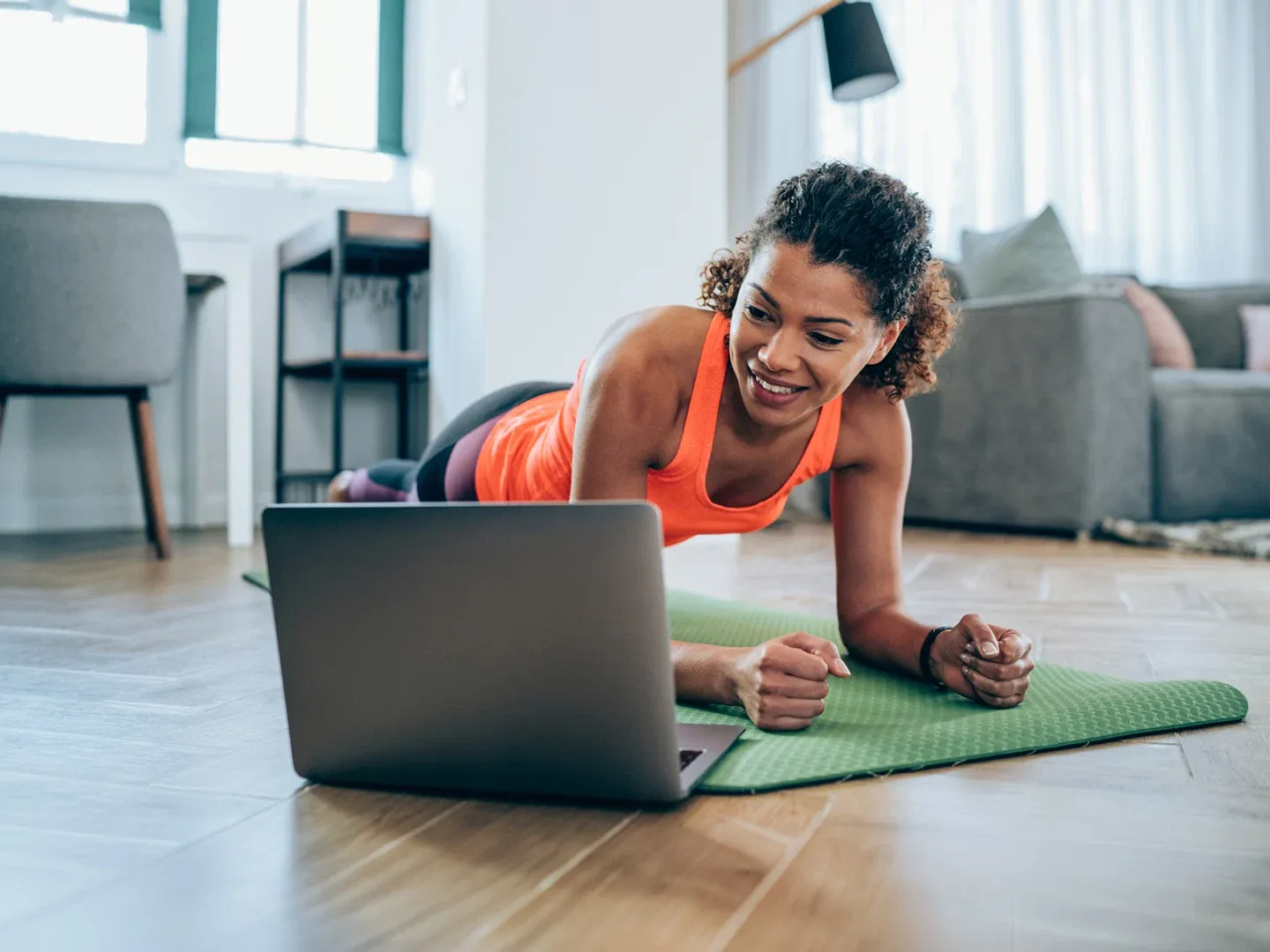A smiling woman holds a forearm plank on a green exercise mat while watching a workout video on her laptop at home.