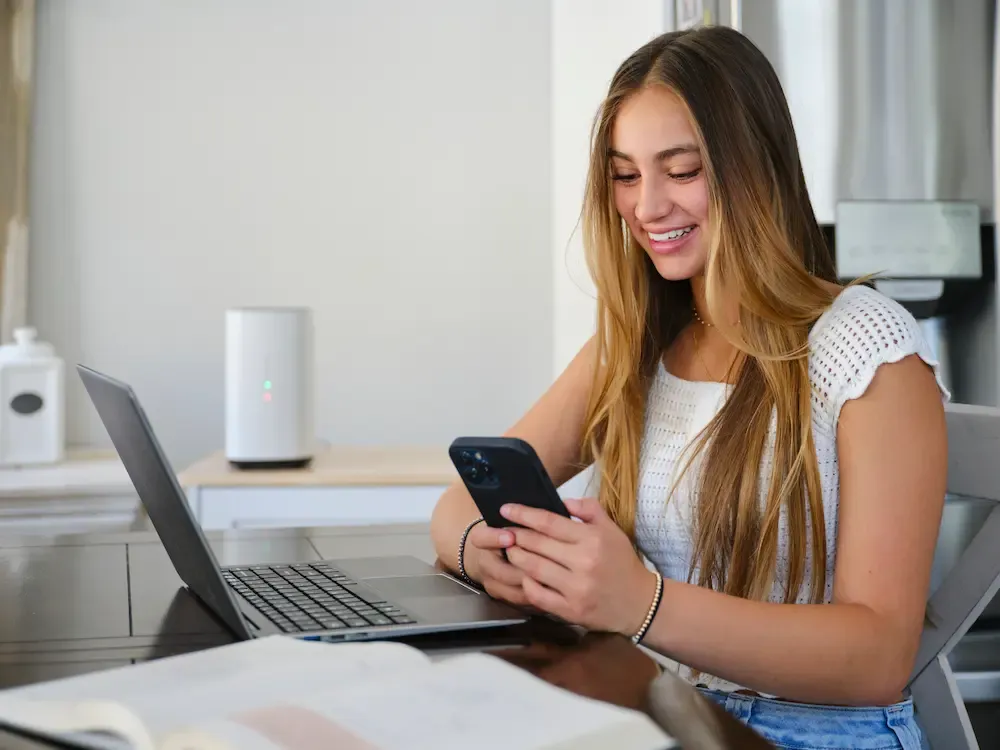 A smiling young woman sitting at a desk with a laptop and open book while using her smartphone, with a white internet router visible in the background.