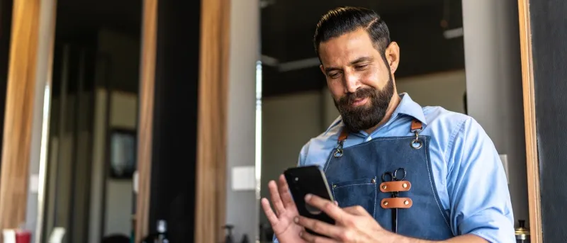 A bearded barber wearing a denim apron checks his smartphone while standing in a salon with large mirrors in the background.