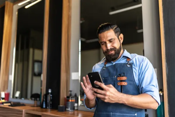 A bearded barber wearing a denim apron checks his smartphone while standing in a salon with large mirrors in the background.
