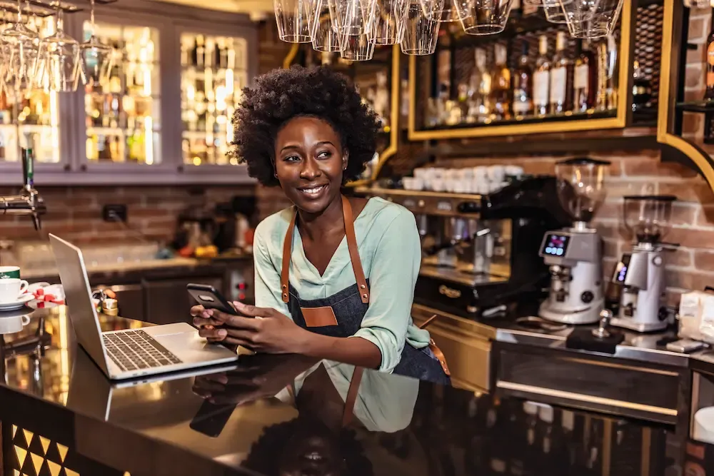 A smiling female barista wearing an apron leans on a counter with a laptop, holding a smartphone in a coffee shop setting.