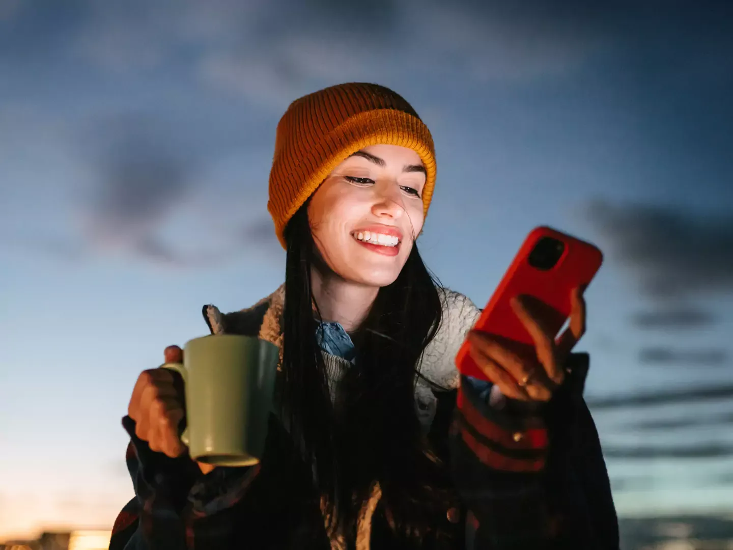 Smiling girl with a beanie and cup of hot cocoa using her phone.