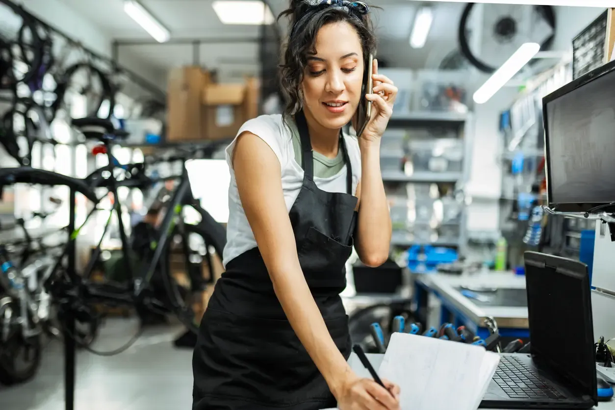 A woman in an apron talks on her phone and takes notes at the counter of a bicycle shop.