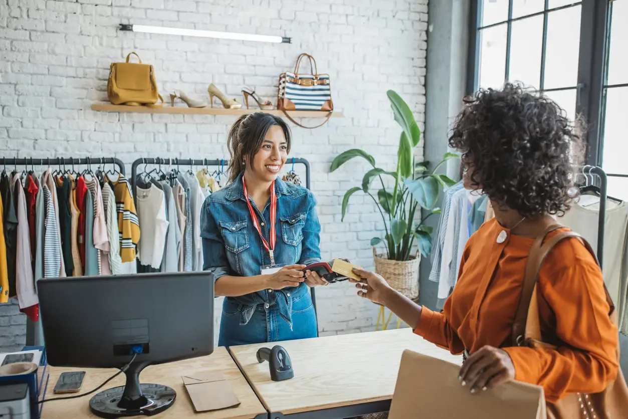 A smiling shop clerk holds a card reader as a customer taps her credit card to pay.