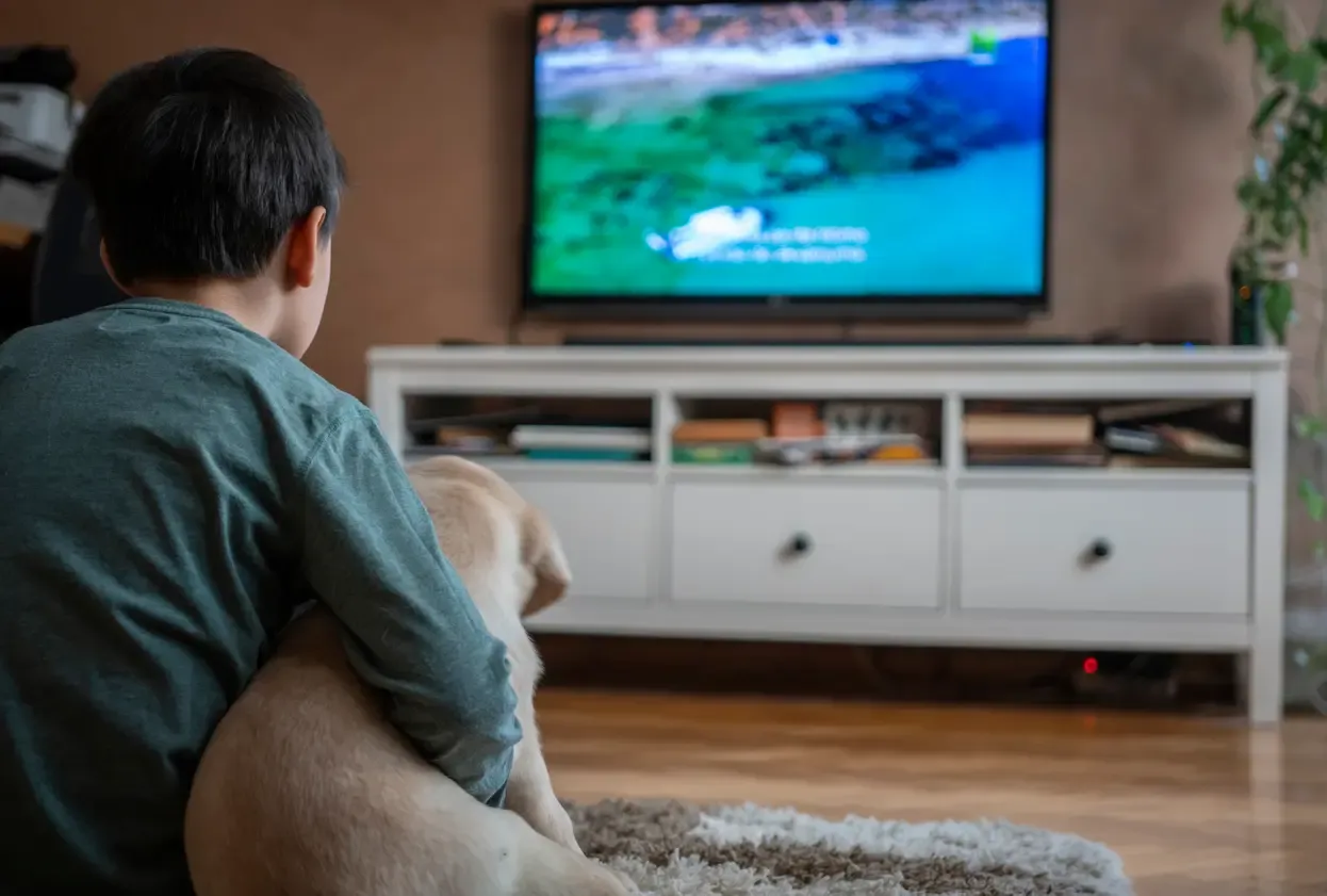 A boy sits on the floor with his arm around a yellow dog, both facing a TV in a living room.