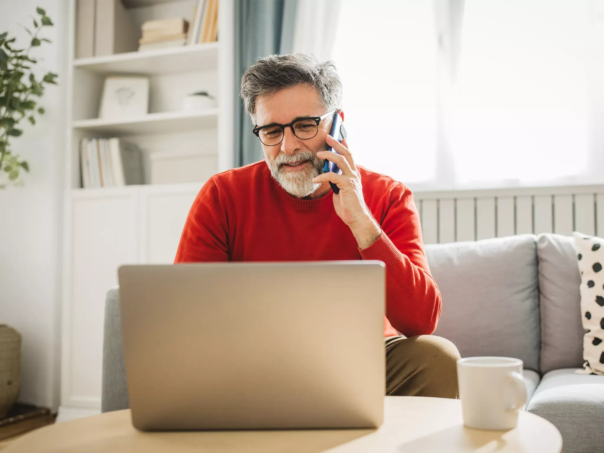 A man in a red sweater chatting on his phone, and using a laptop in his home.