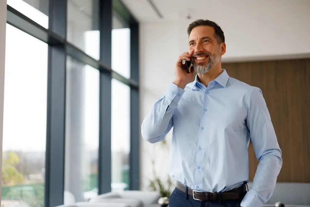 A smiling businessman in a blue dress shirt stands in a bright, modern office while talking on a smartphone near a large window.