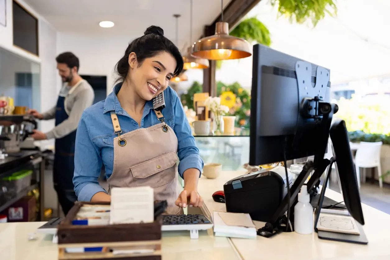 A smiling female cafe worker wearing an apron takes an order over the phone while typing on a computer at the service counter.
