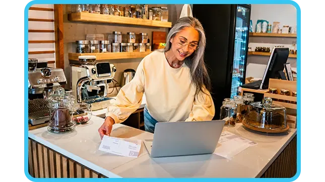 A smiling middle-aged woman with long gray hair manages shop finances using a laptop and paperwork on a cafe counter.