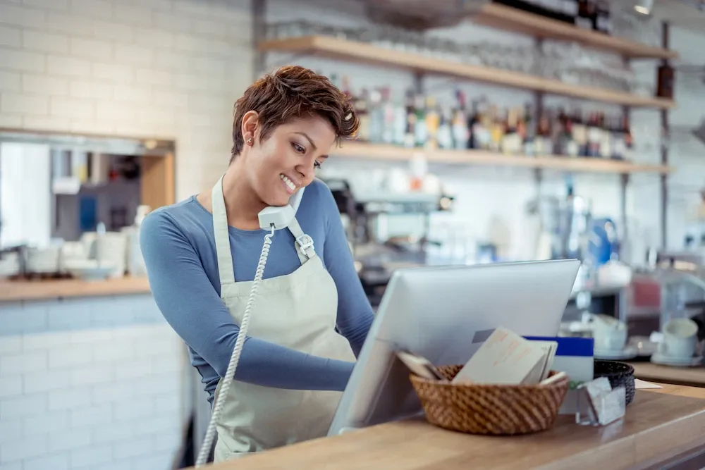 A smiling woman with short hair wearing an apron and blue shirt is talking on a landline phone while looking at a computer monitor behind a cafe counter.