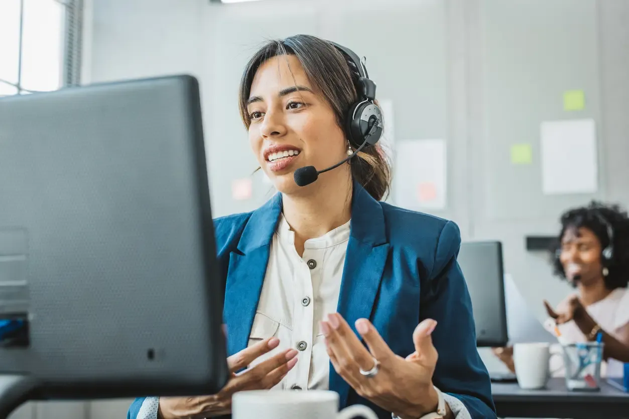 A smiling woman wearing a headset speaks to a customer at her computer in a call center.