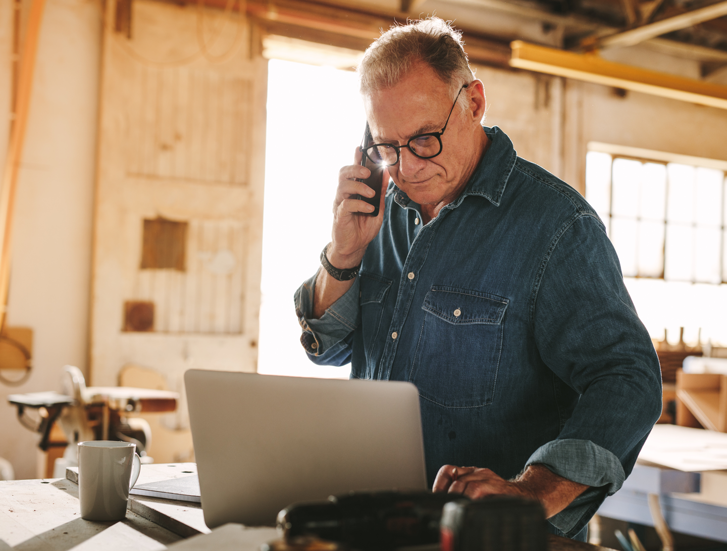 carpenter in his shop using the phone and looking at his laptop