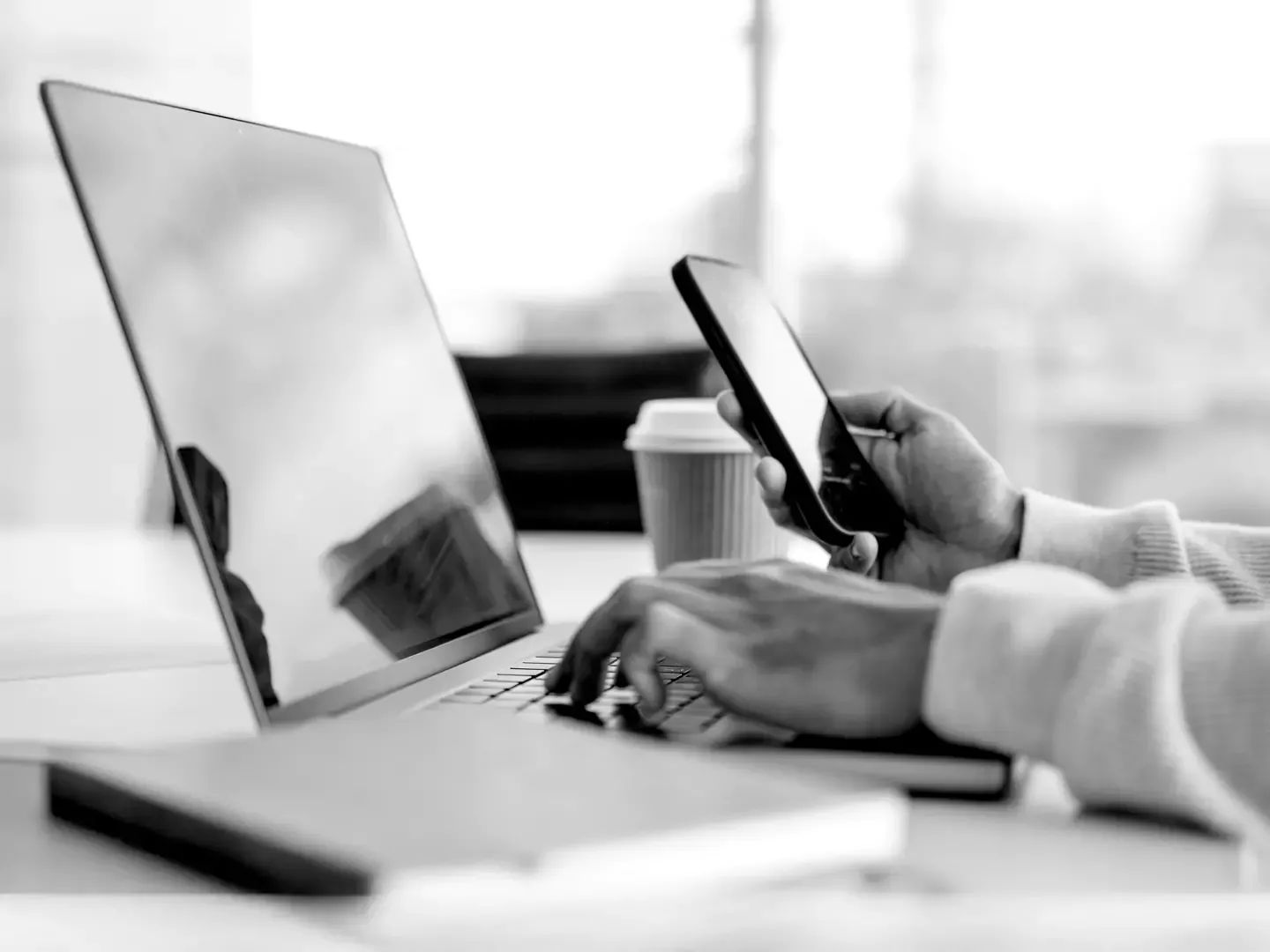 Close up hands working at a keyboard and using a mobile device, with a coffee cup in the back.