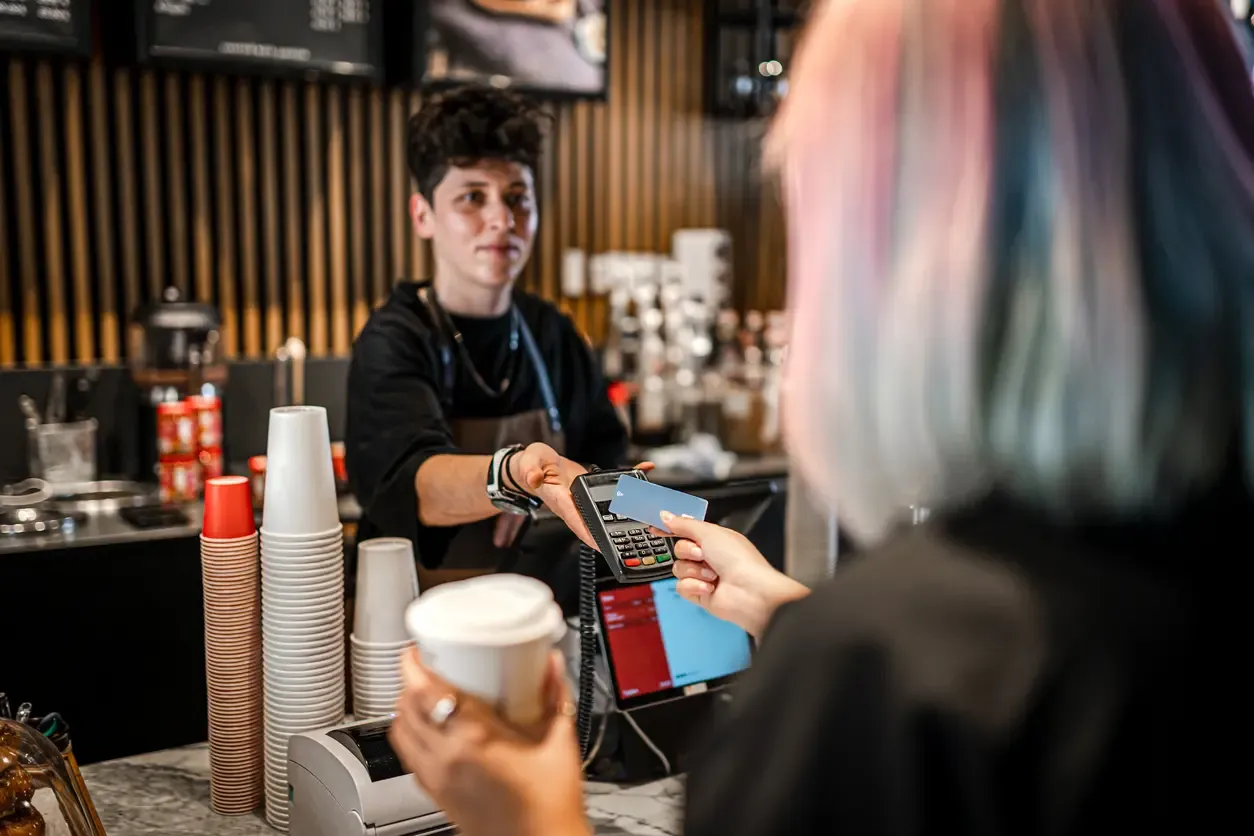 A barista holds a card reader as a customer taps a credit card to pay at a coffee shop counter.