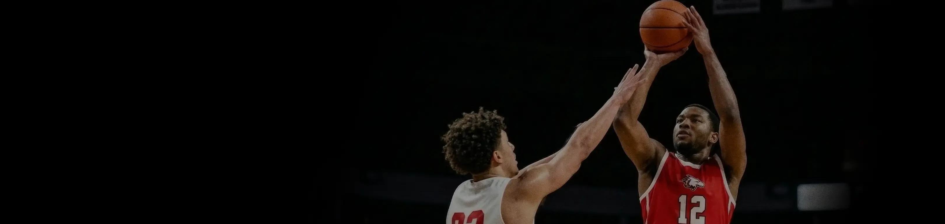 A basketball player in a red jersey jumps to take a shot while a defender in a white jersey reaches out to block him against a dark background.
