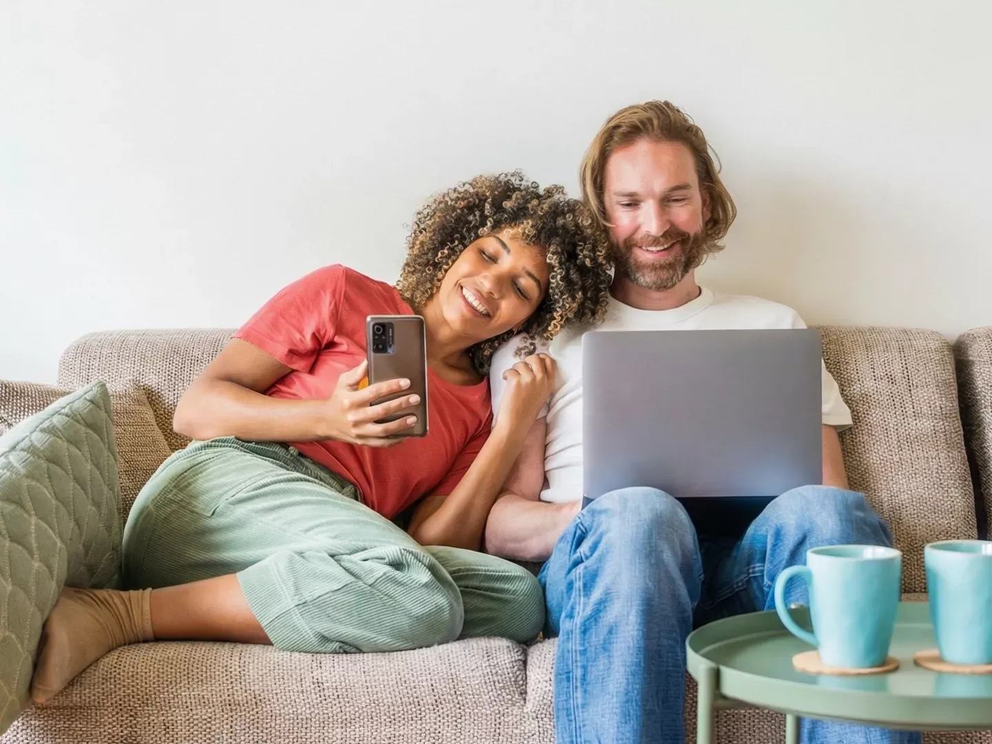 A smiling couple sits on a couch together while the woman looks at her smartphone and the man works on a laptop.