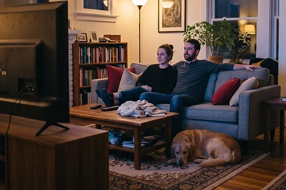 A couple relaxes on a sofa watching TV while their dog rests nearby.