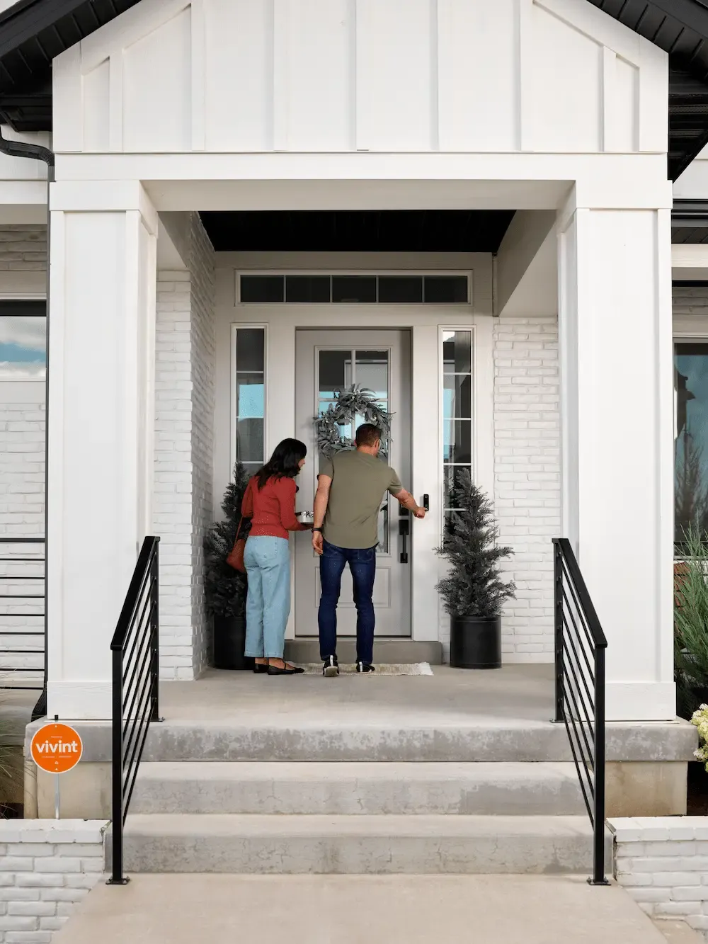 A man and woman stand on the porch of a modern white house as the man reaches for a smart lock and doorbell.