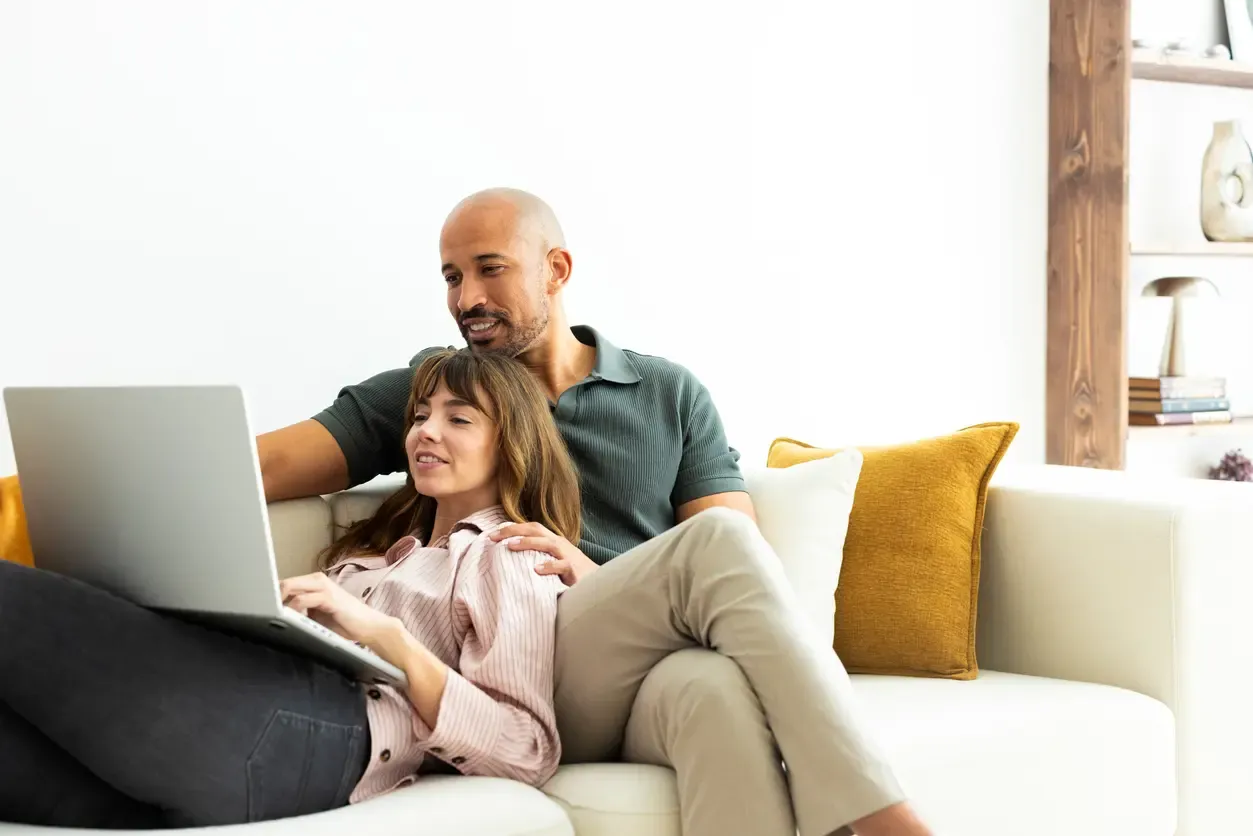 A smiling couple relaxes on a white couch browsing a laptop together at home.