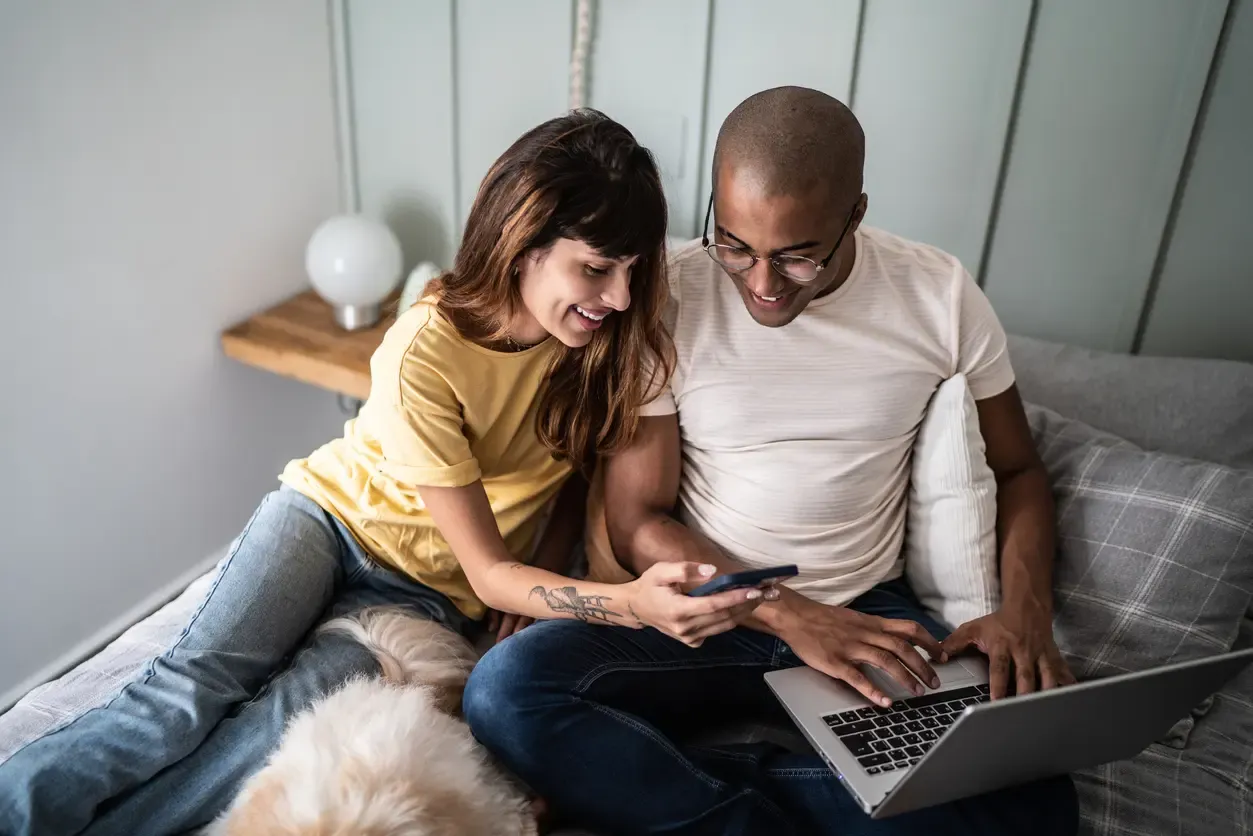 A smiling couple sits on a bed sharing a laptop and smartphone with their dog nearby.