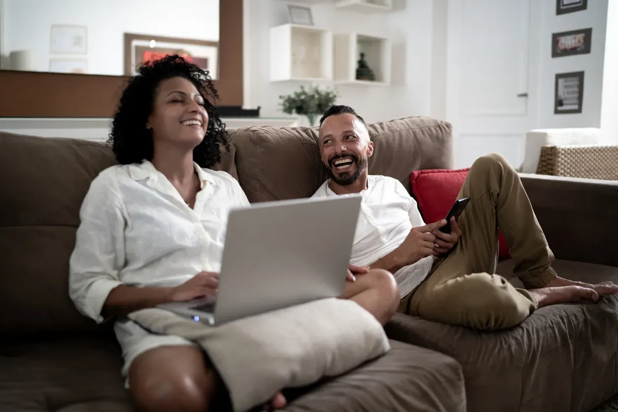 A man and woman laugh together on a couch while she uses a laptop.