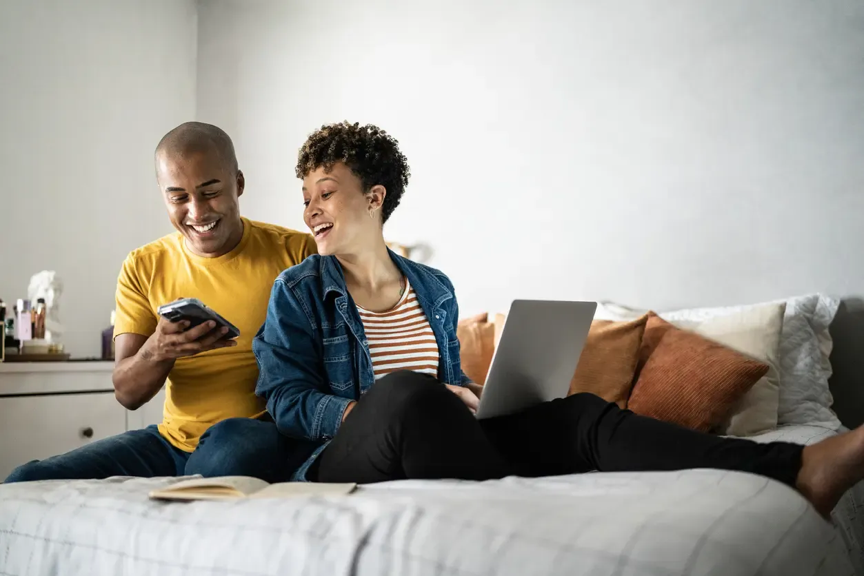 A happy young couple sits together on a bed, laughing as they look at a smartphone and use a laptop computer.