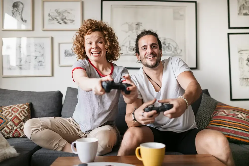 An excited man and woman sitting on a gray sofa holding controllers while playing video games together in a decorated living room.