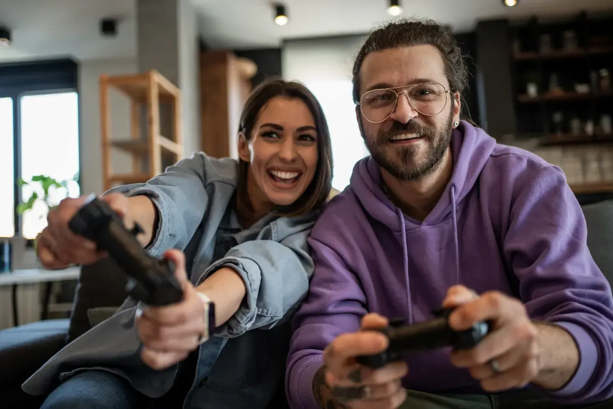 A smiling young couple sitting on a couch and holding controllers while playing a video game together.
