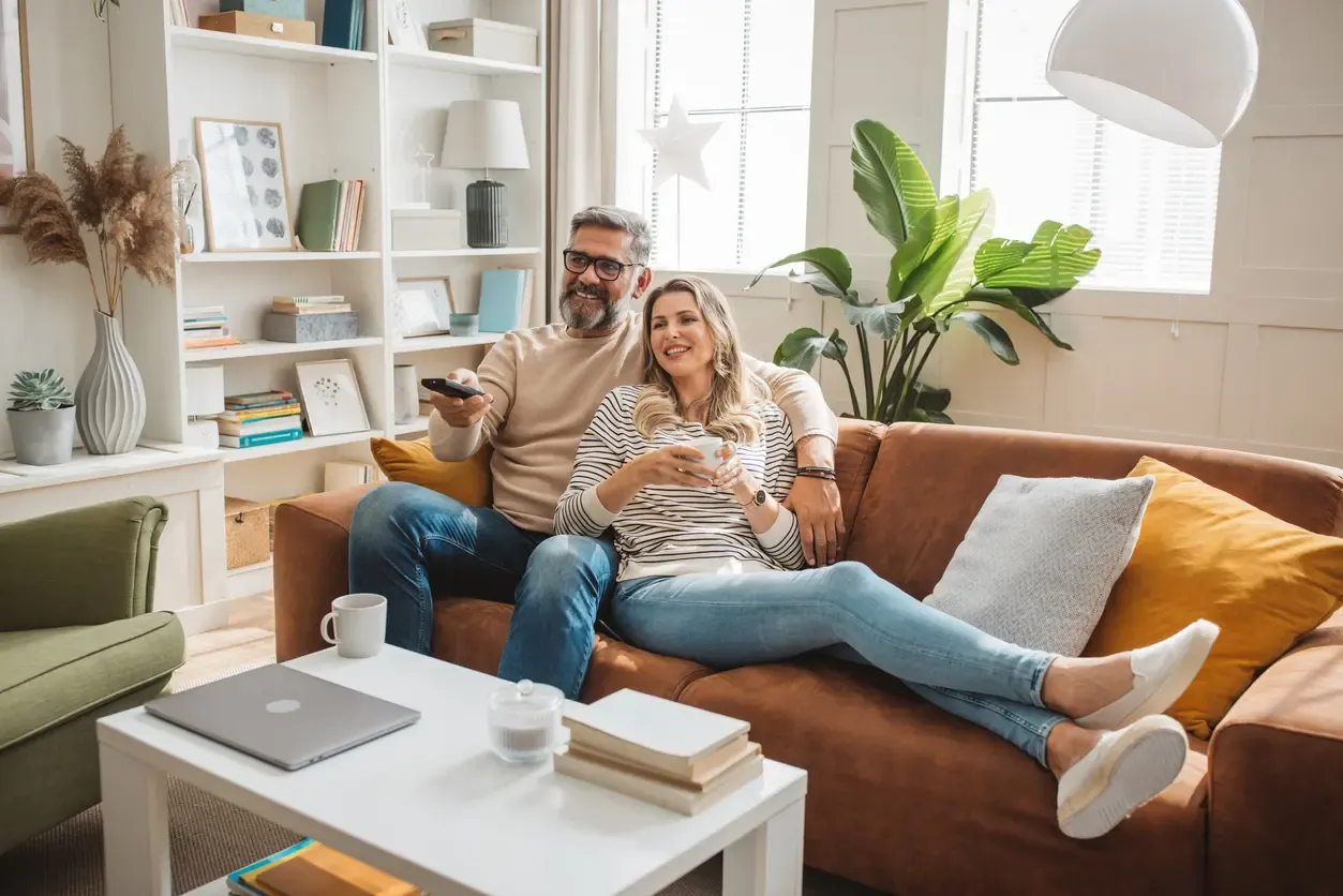 A smiling couple relaxes together on a brown sofa, the man holding a remote control and the woman cradling a mug.