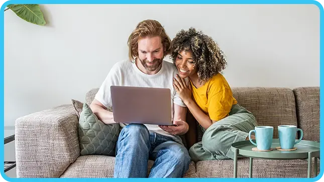 A smiling couple sits close together on a couch, looking at a laptop in a bright living room.